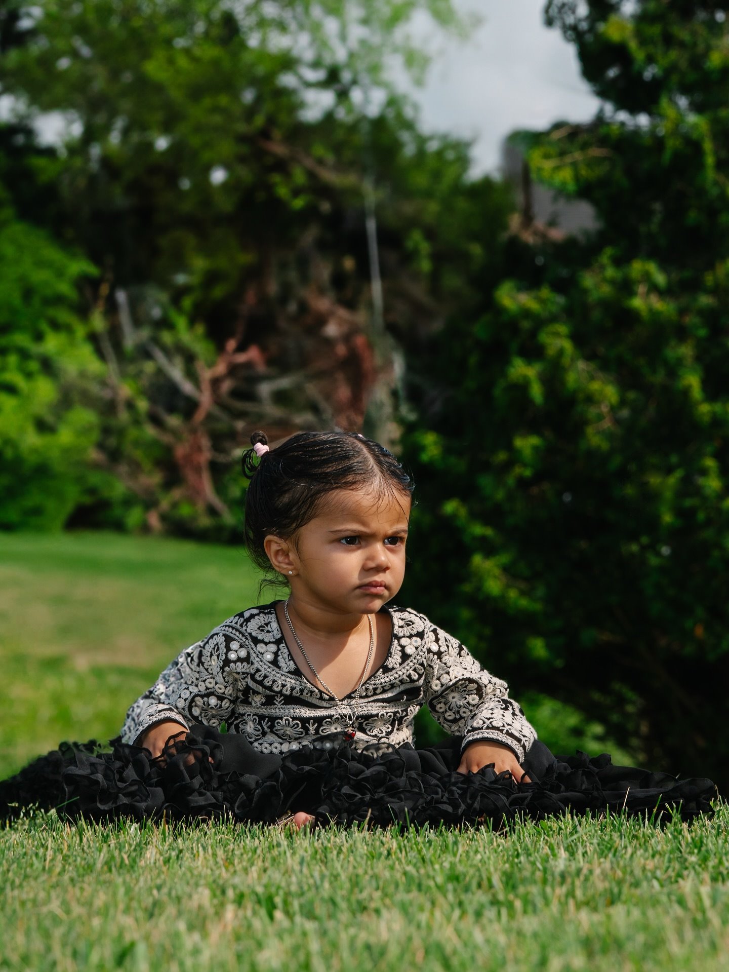 A toddler in a black embroidered dress sitting on the grass with a serious look