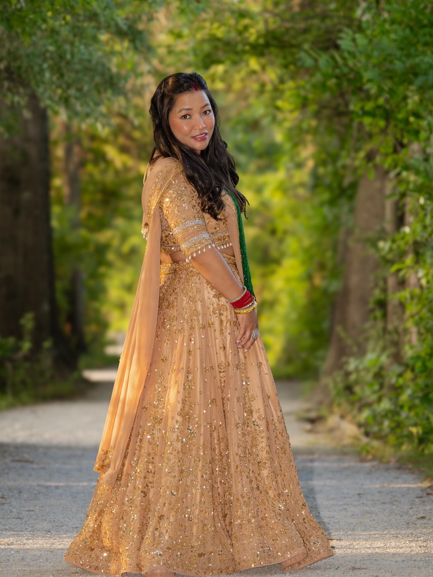A bride in a gold lehenga looking back over her shoulder on a tree-lined path