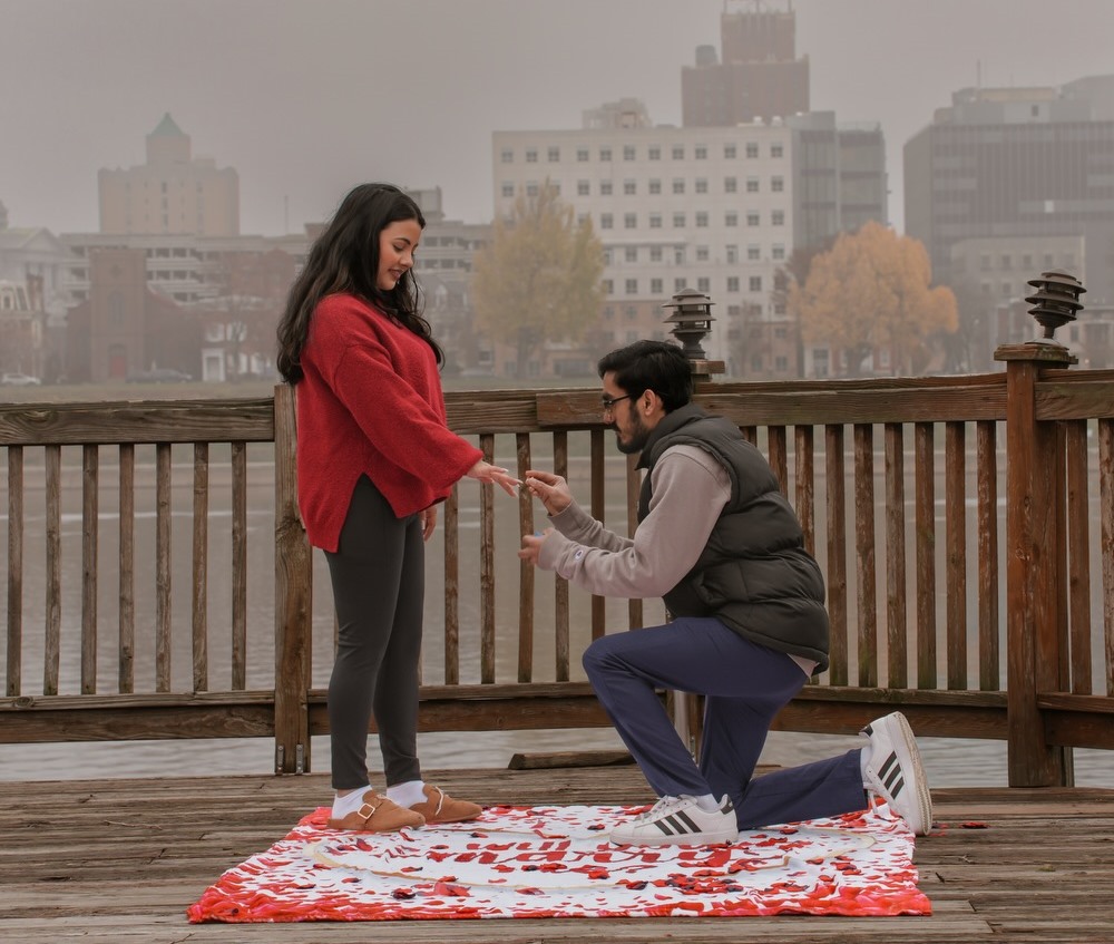 A man on one knee proposing on a wooden deck by the river on a foggy morning, rose petals at their feet